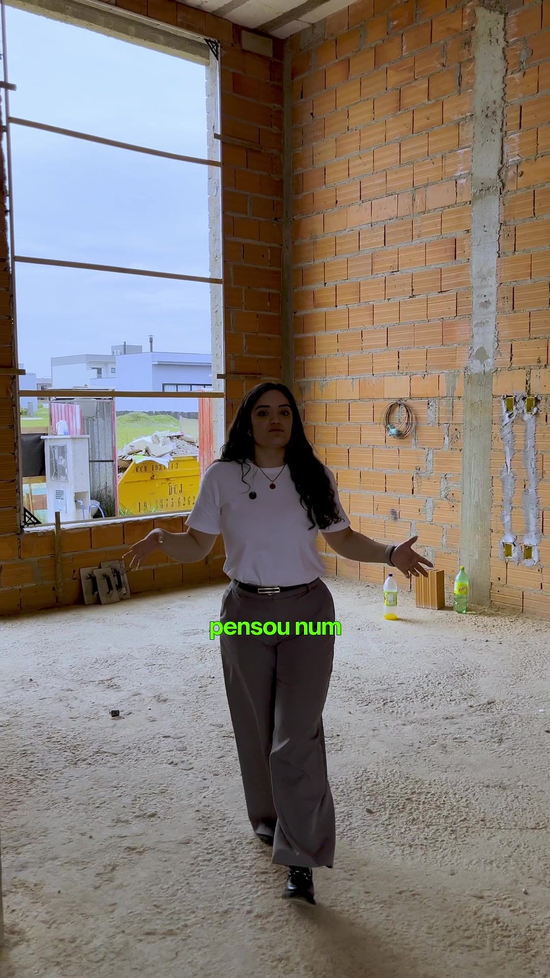 Mulher de camiseta branca falando para a câmera dentro de uma casa em obra, com paredes de tijolo e uma janela alta ao fundo